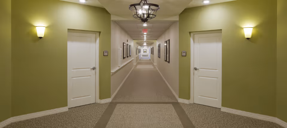 Long interior hallway with green accent walls, white doors on both sides, wall sconces and framed artwork leading to a distant exit.