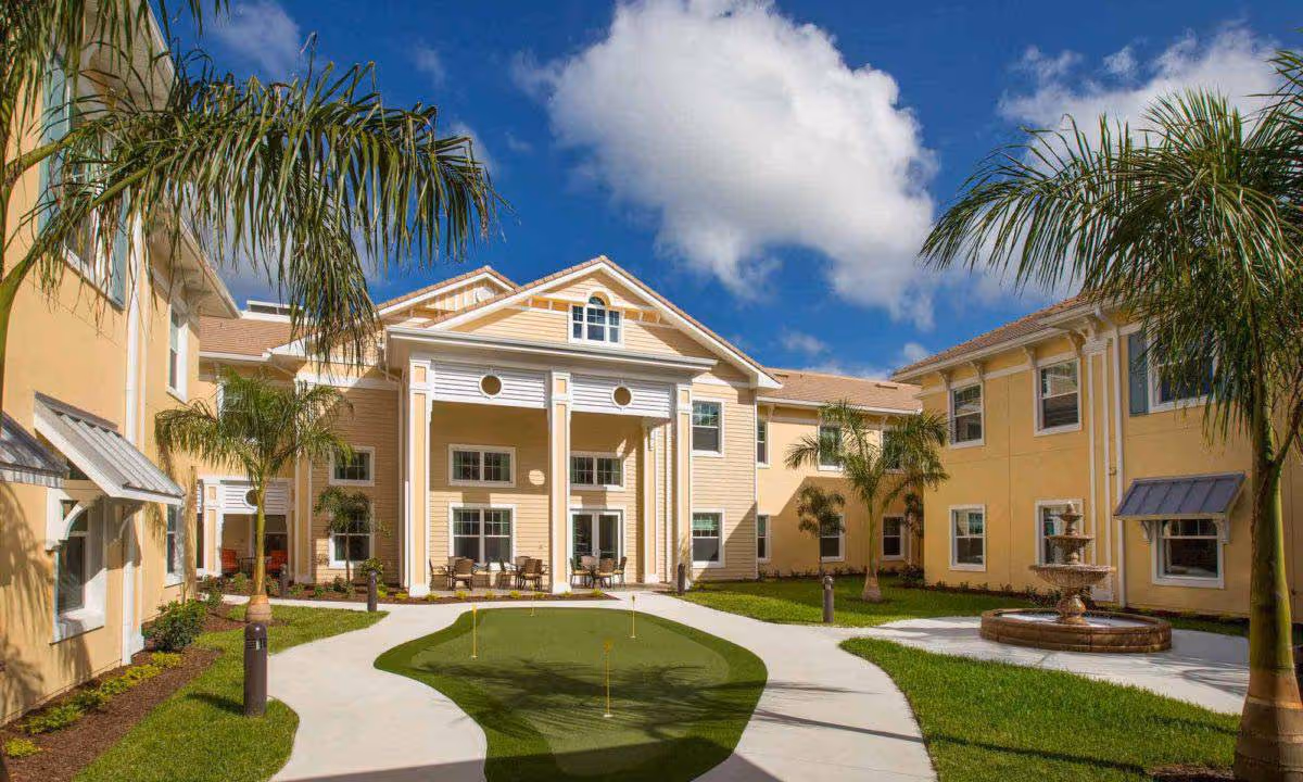 Sunny courtyard with palm trees, a putting green and fountain in front of a yellow two-story senior living building.