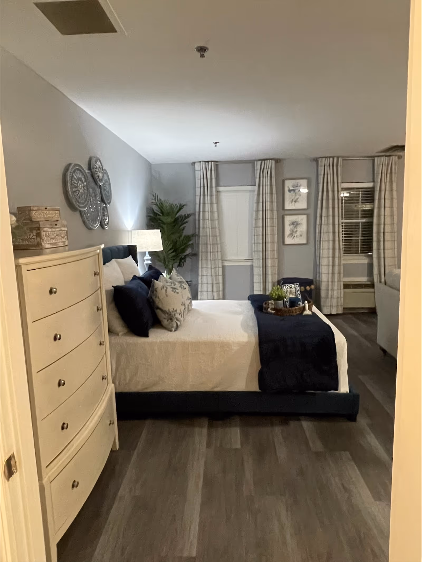 A neatly made bed with white bedding and dark blue pillows and throw blanket in a bedroom. There is a white chest of drawers on the left side, a lamp, decorative wall plates, and a potted plant. Two windows with plaid curtains and framed artwork are visible on the far wall.