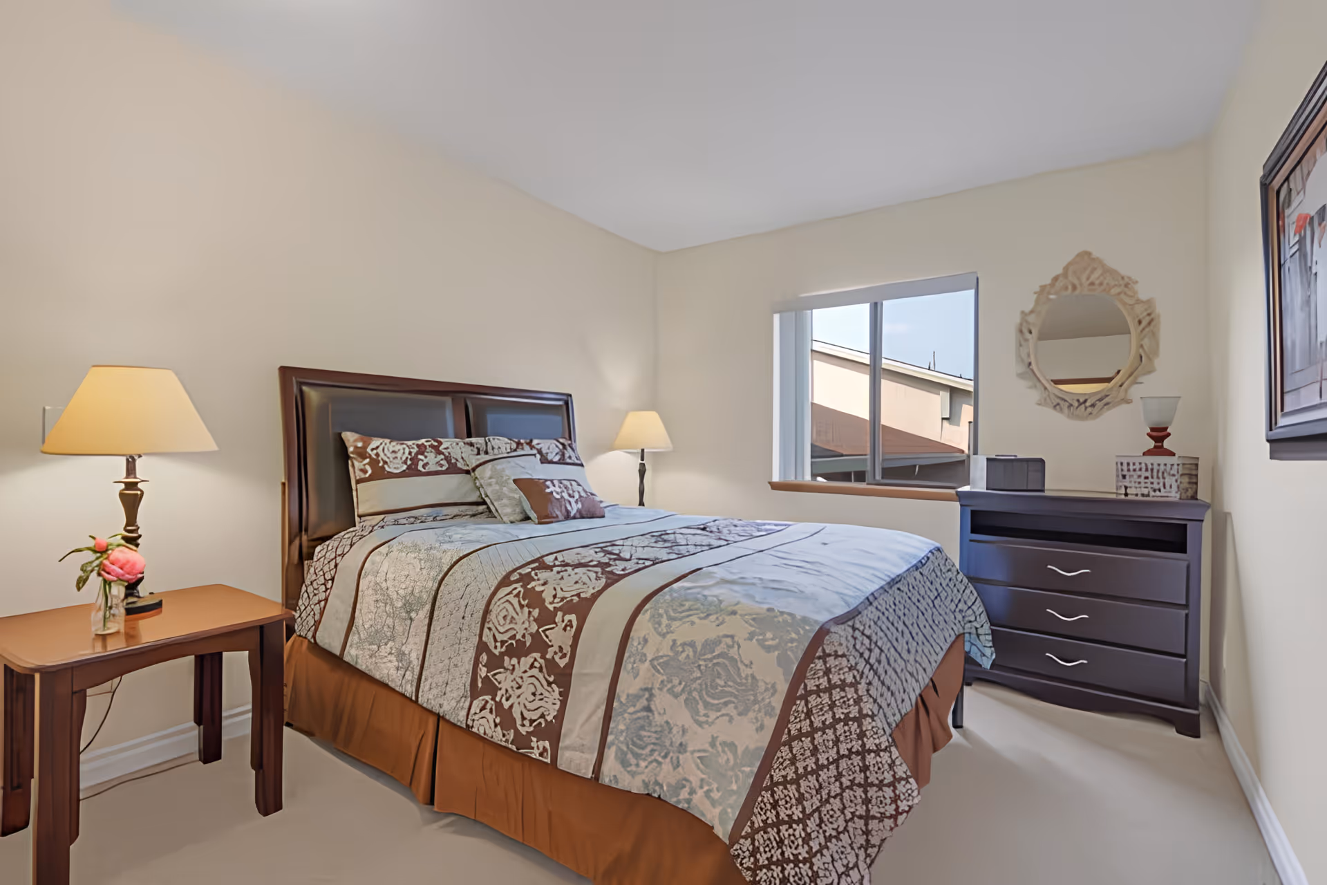 A neatly made bed with a patterned bedspread and multiple pillows in a bedroom. There are two bedside tables with lamps on either side of the bed. A window with a view of an adjacent building is on the right wall. A dark wooden dresser with a decorative mirror and some items on top is also visible. The walls are painted light beige.