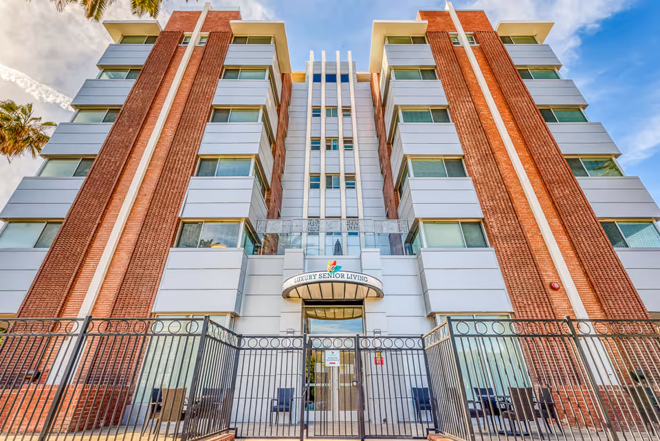 Front exterior view of a multi-story senior living facility building with a gated entrance. The building features red brick and white paneling with multiple windows and a sign above the entrance that reads 'Luxury Senior Living'. Palm trees are visible on the sides under a partly cloudy sky.