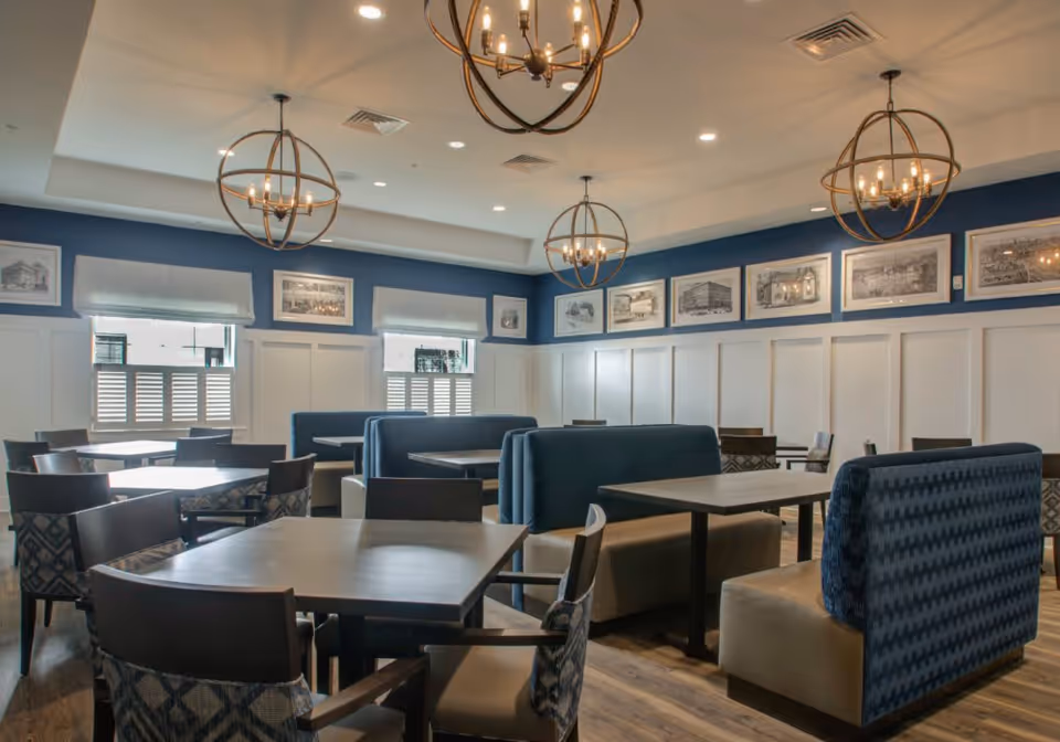 A dining room with multiple tables and chairs, featuring blue upholstered booth seating and wooden chairs with patterned cushions. The walls are decorated with framed black and white pictures, and there are three spherical chandeliers hanging from the ceiling. The room has large windows with white shutters and a blue accent wall.