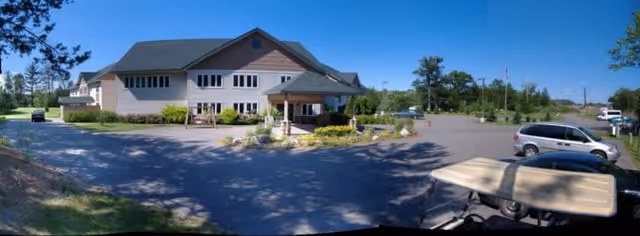 Wide panoramic view of a two-story assisted living facility building with a covered entrance, surrounded by a parking lot with several parked cars. The building is light-colored with multiple windows and a gabled roof. Trees and greenery are visible around the property under a clear blue sky.