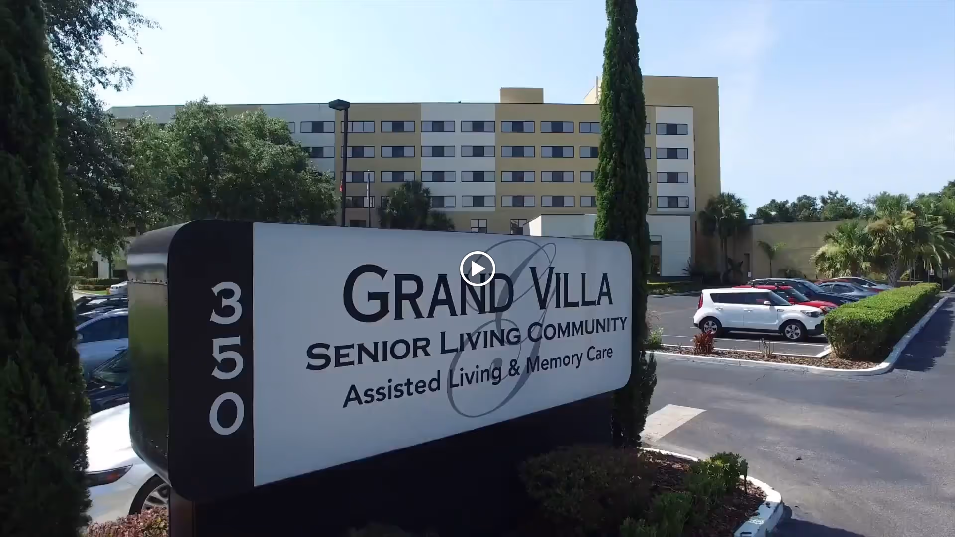 Entrance sign reading 'Grand Villa Senior Living Community' with the facility building and parking lot behind it.
