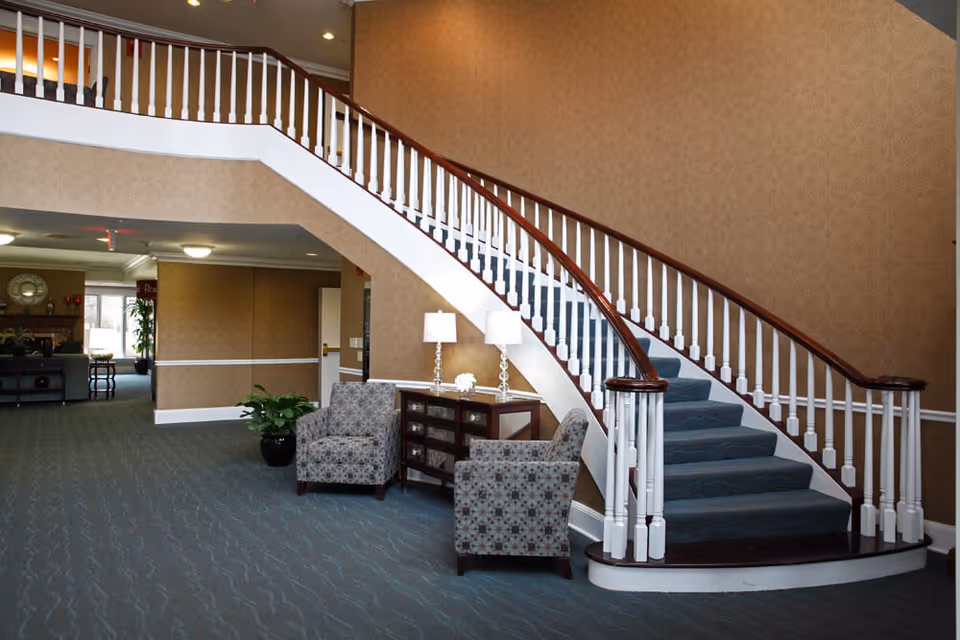Interior view of a senior living facility lobby area featuring a curved staircase with white railings and dark wooden handrails. Two patterned armchairs are placed around a small wooden table with two lamps and a decorative item. The walls are covered with beige wallpaper, and the floor is carpeted in blue. In the background, there is a hallway leading to other rooms with additional seating and plants.