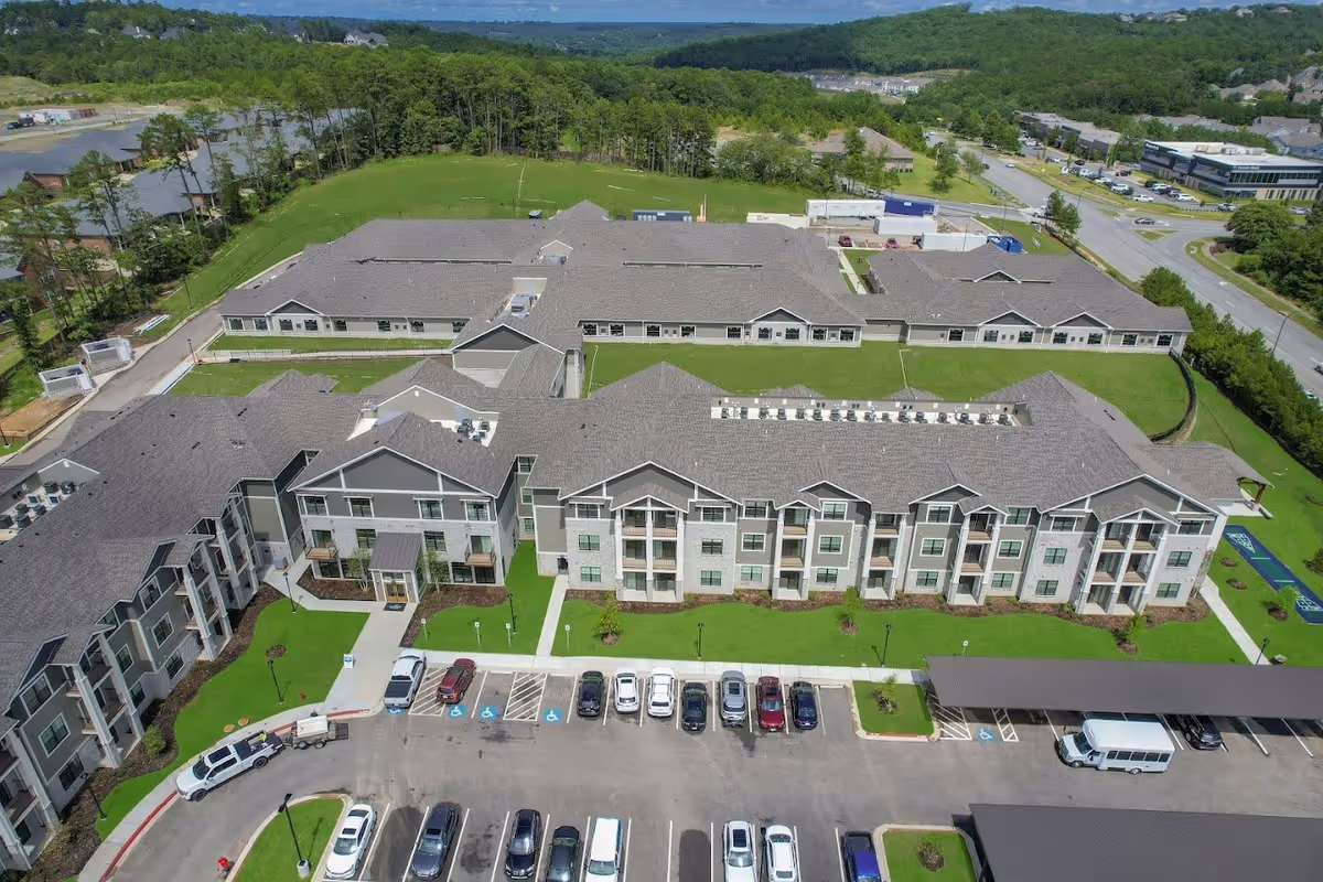 Aerial view of a large retirement community building with multiple wings, surrounded by green lawns and trees. There is a parking lot with several cars and a covered parking area in front of the building. Roads and additional buildings are visible in the background.