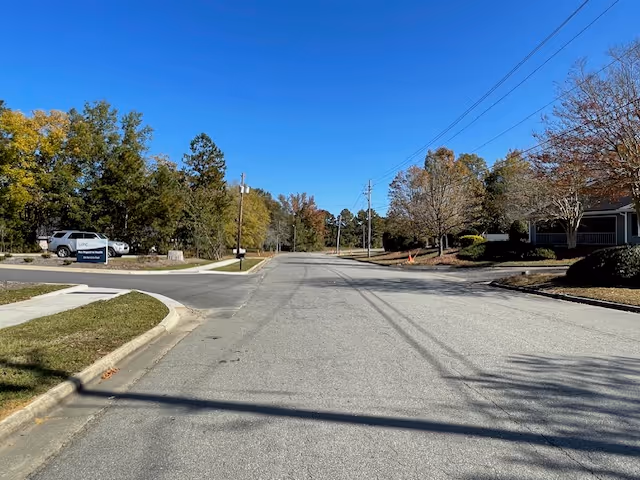 A wide paved road lined with trees on both sides under a clear blue sky. There is a white vehicle parked on the left side near a sign, and a house partially visible on the right side. Power lines run along the road.