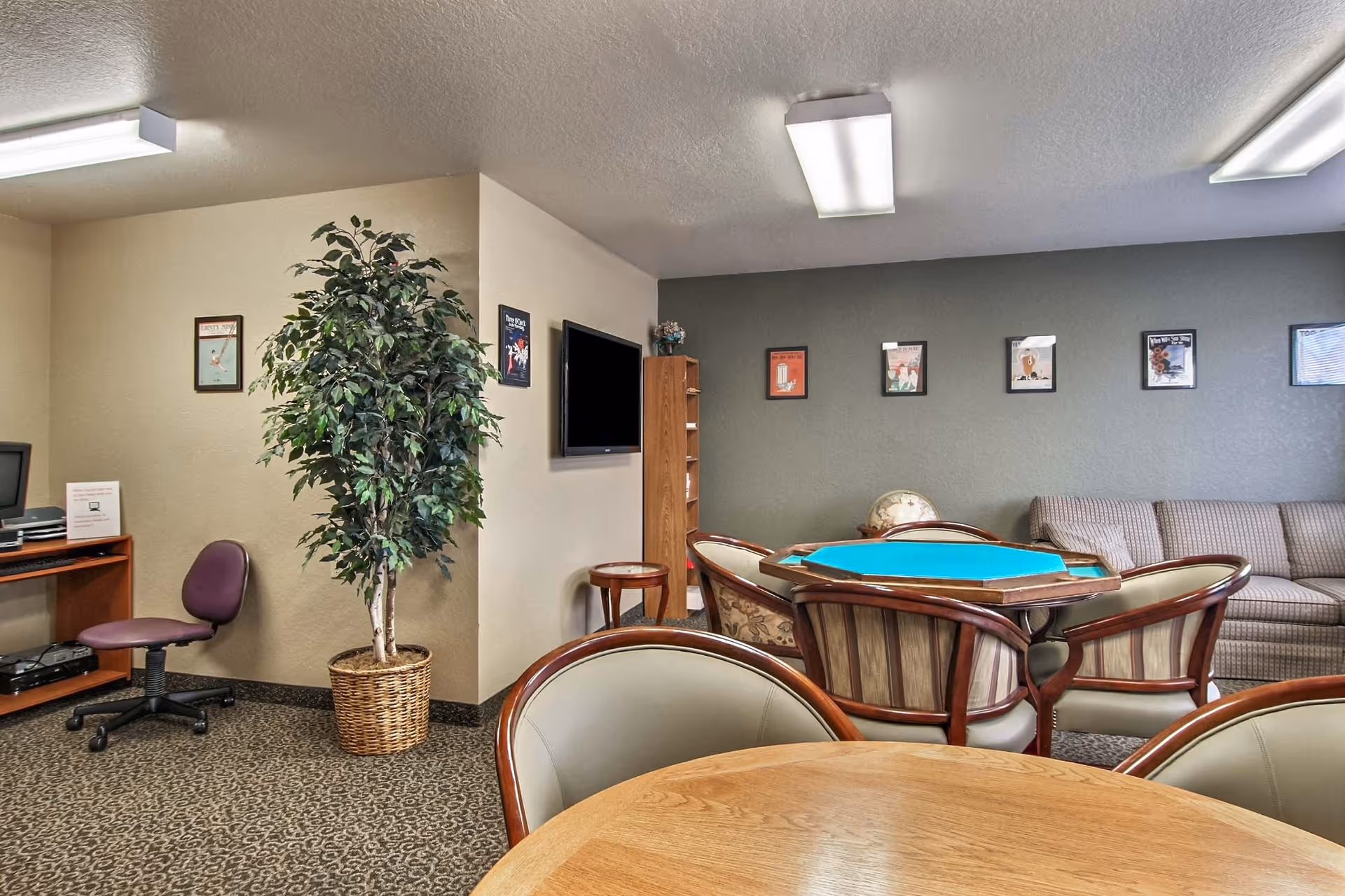 Communal lounge with a card table and chairs, sofa, wall-mounted TV, and a potted plant in a senior living facility.