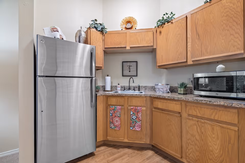 A small kitchen area with wooden cabinets, a stainless steel refrigerator, a microwave on the countertop, a sink with a faucet, and decorative items including a silver vase, a flower-shaped decoration, and two colorful towels hanging on the cabinet doors.