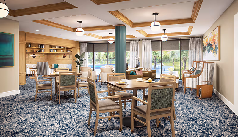 A spacious and well-lit common area in a senior living facility with multiple wooden tables and chairs arranged on a patterned blue carpet. The room features large windows with views of greenery outside, a teal column in the center, ceiling lights, and comfortable seating areas with sofas and armchairs. There are paintings on the walls and a bookshelf in the background.