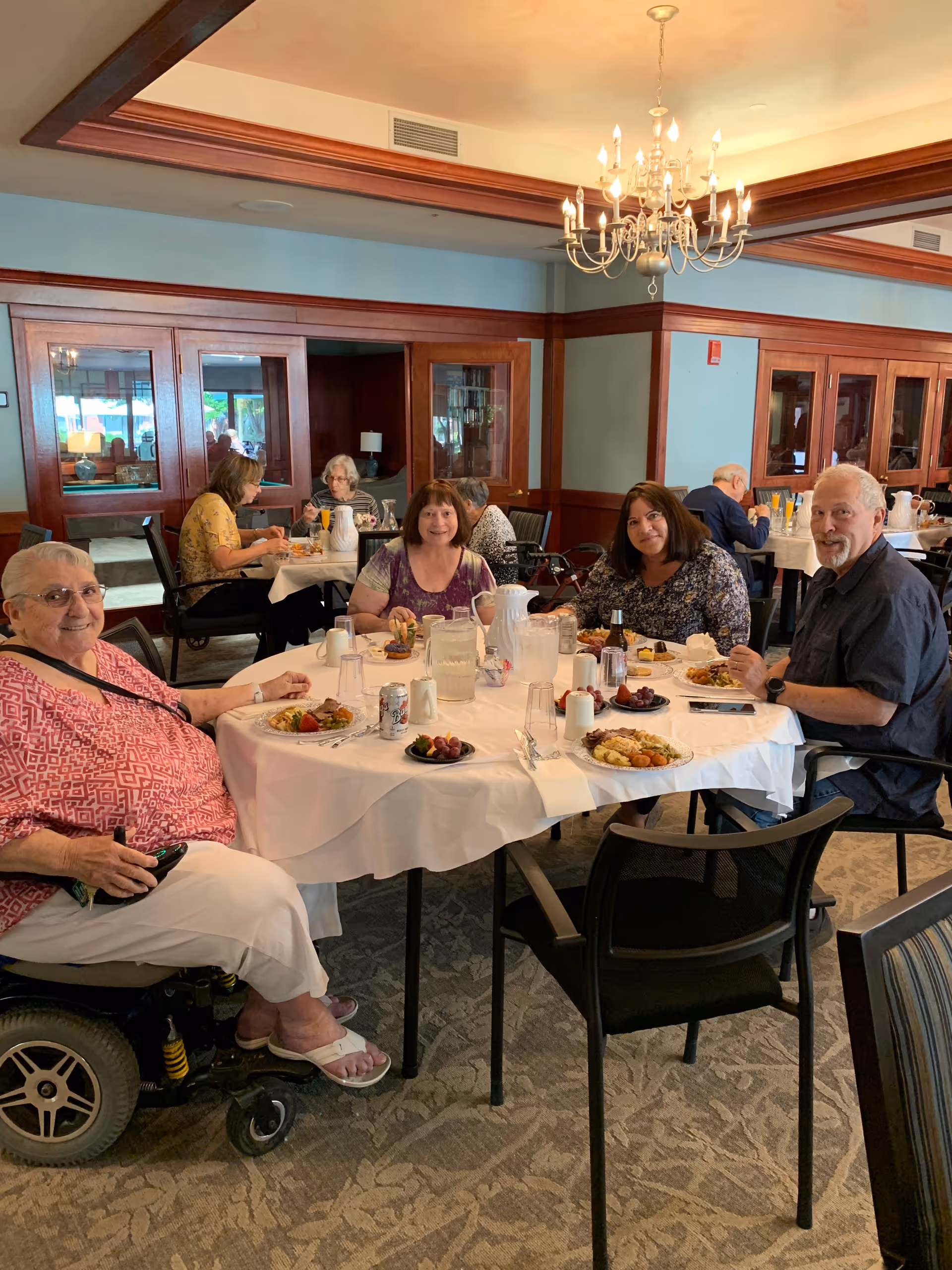 Four elderly adults sitting around a round dining table in a well-lit dining room, enjoying a meal together. The table is covered with a white tablecloth and has plates of food, pitchers of water, and drinks. The room features wooden paneling, a chandelier, and other diners seated at tables in the background.