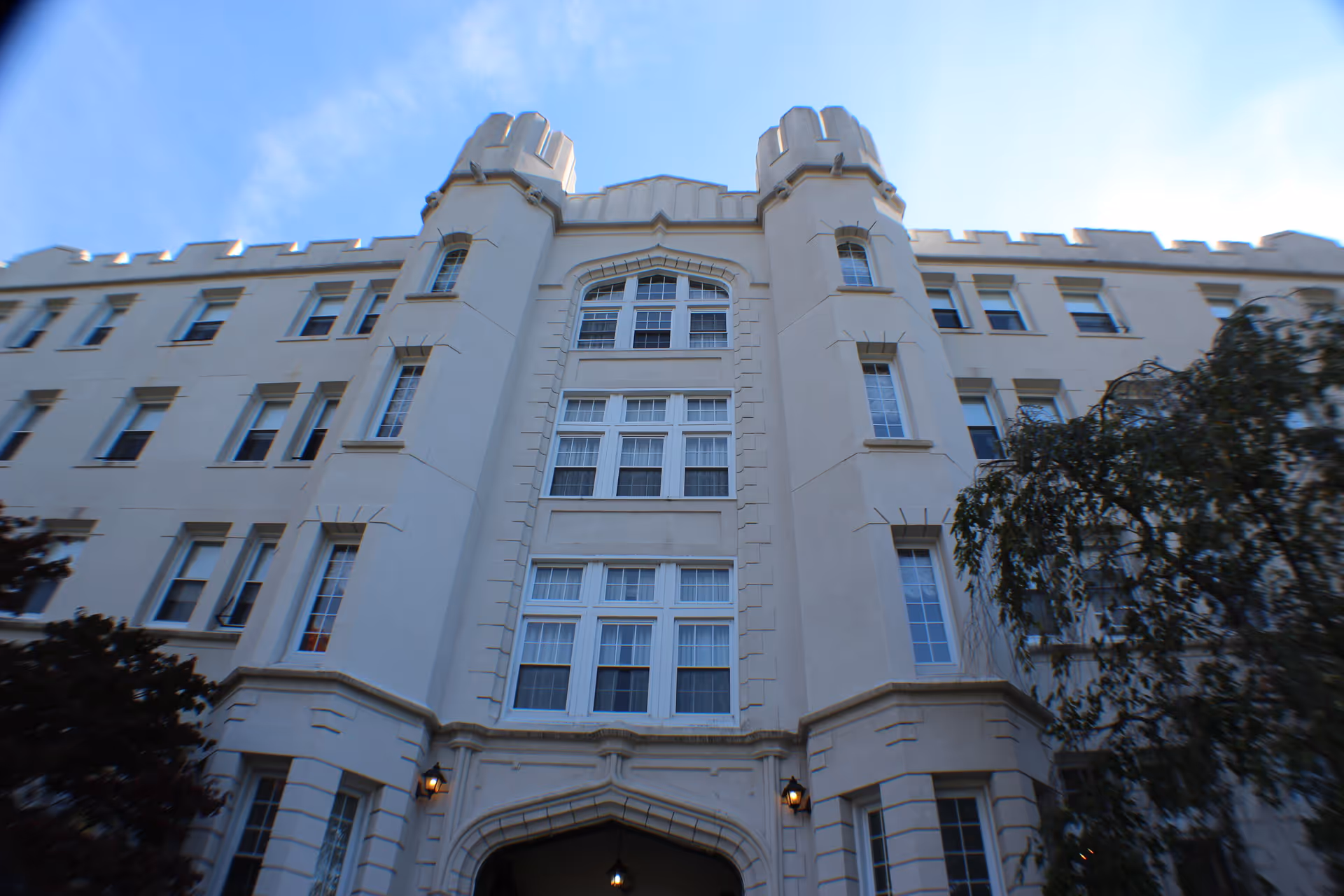 Exterior view of a large, multi-story building with castle-like architectural features including turrets and crenellations. The building is light-colored with many windows and an arched entrance. Trees partially frame the building on either side under a blue sky.