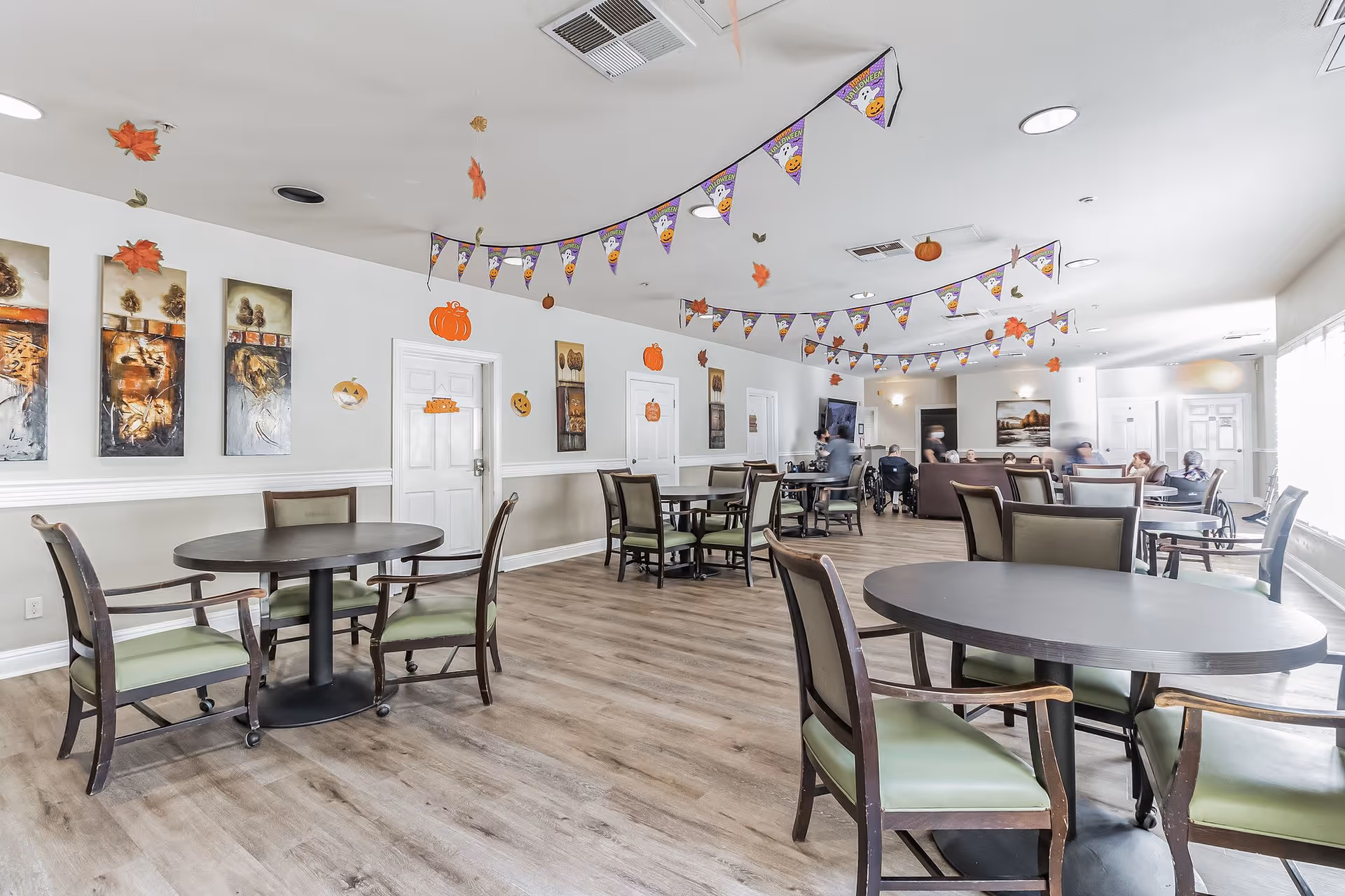A spacious common area in a senior living facility decorated with Halloween-themed hanging decorations including pumpkins, leaves, and banners. The room has several round tables with green cushioned chairs, wood flooring, and white walls adorned with artwork. In the background, a few seniors are seated on couches and chairs near a television.