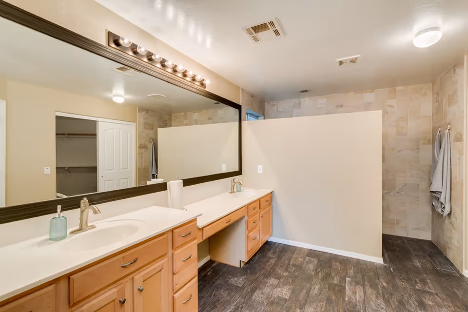 A spacious bathroom with a large mirror above a double sink vanity with wooden cabinets. The floor has dark wood-like tiles, and there is a walk-in shower area with beige tiled walls partially visible behind a half wall. Two gray towels hang on the wall near the shower.