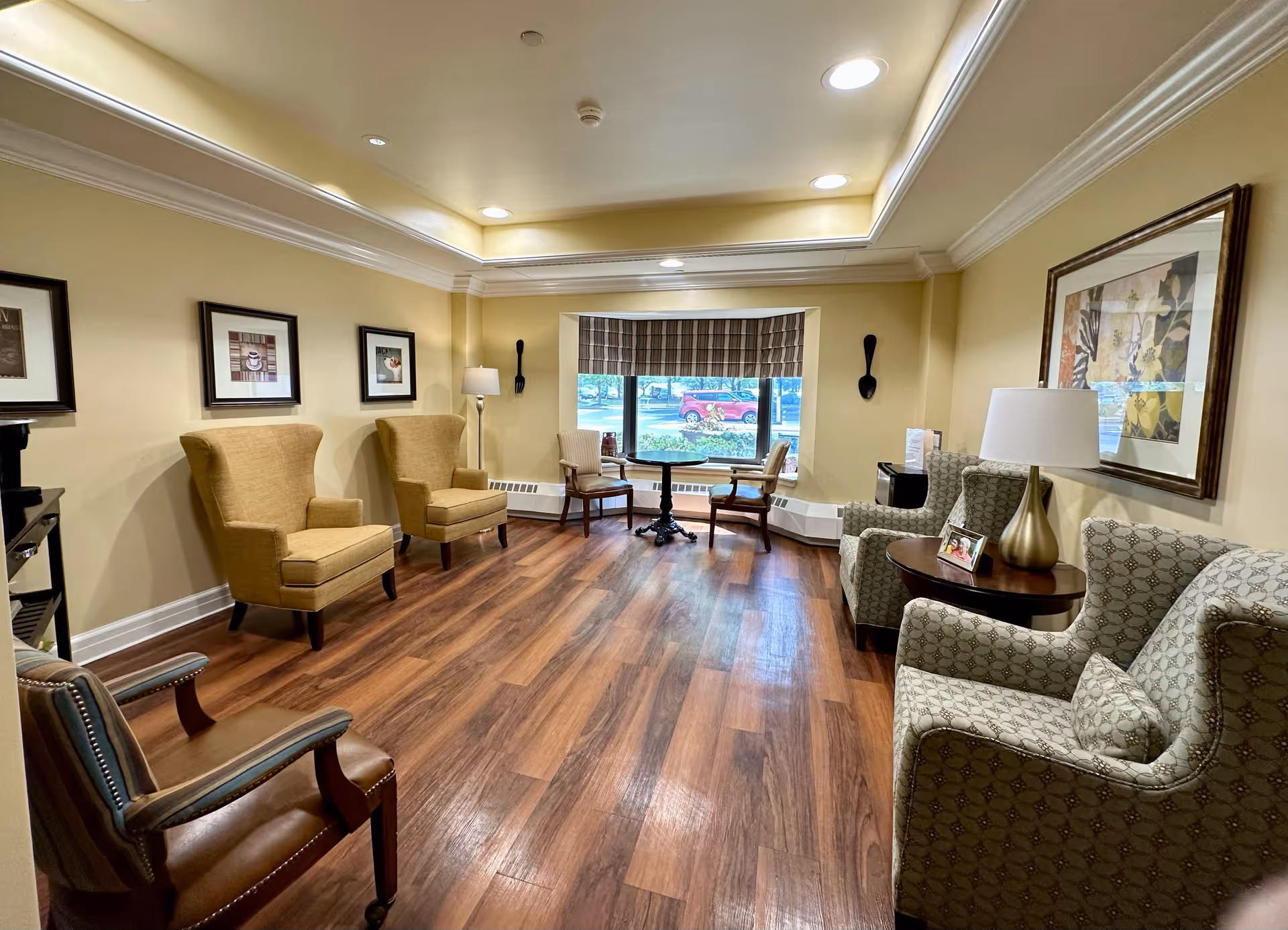 A cozy sitting area in a retirement facility with several upholstered armchairs arranged around the room. The space features wood flooring, beige walls with crown molding, framed artwork, a window with a plaid valance, and a small round table with two chairs near the window. A table lamp and a small photo frame are on a side table between two armchairs.