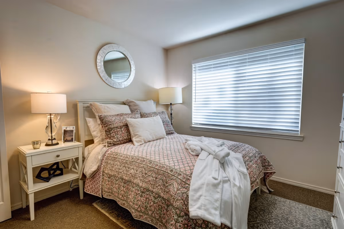 A cozy bedroom with a neatly made bed featuring patterned bedding and multiple pillows. A white robe is draped over the foot of the bed. There is a white nightstand with a lamp, a small candle, and a framed photo on the left side of the bed. A round mirror hangs on the wall above the headboard. A large window with closed blinds is on the right side, allowing soft natural light into the room. The floor is carpeted with a patterned rug partially visible under the bed.