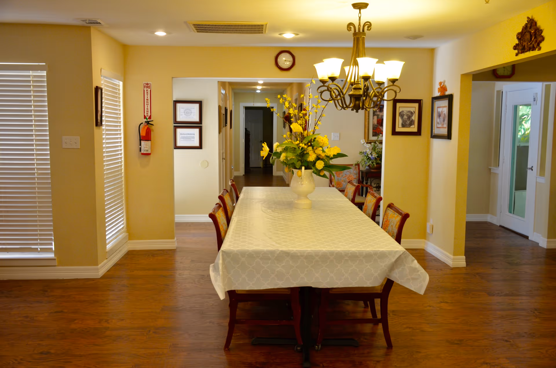 A dining room with a long table covered by a white patterned tablecloth and surrounded by wooden chairs with floral cushions. A large vase with yellow flowers is centered on the table. The room has wooden flooring, beige walls, framed pictures on the walls, a chandelier with multiple lights hanging above the table, and windows with white blinds on the left side.