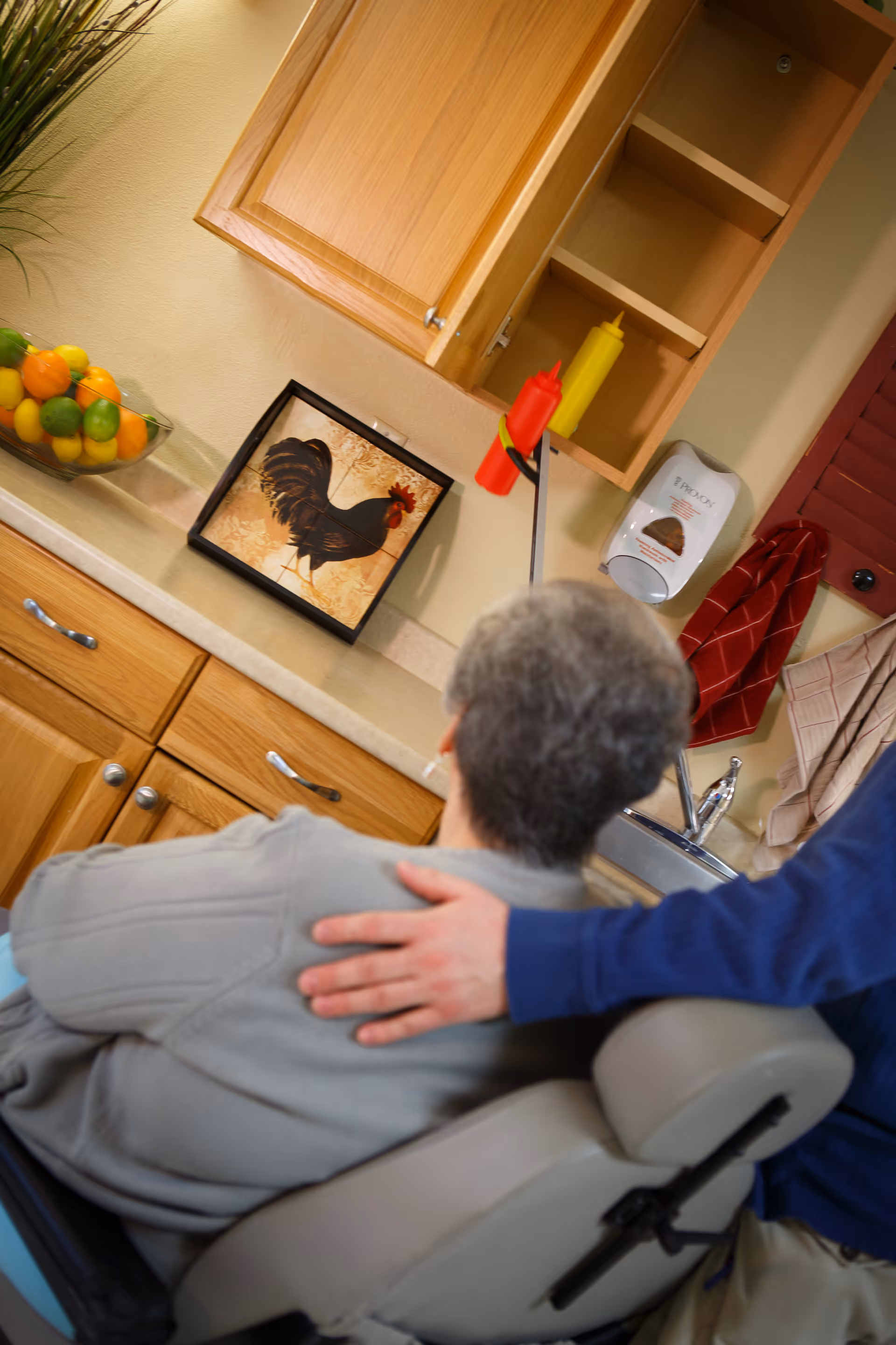 An elderly person sitting in a chair facing a kitchen counter with wooden cabinets. A hand rests gently on the person's shoulder. On the counter, there is a framed picture of a rooster, a bowl of assorted citrus fruits, and condiment bottles. A soap dispenser and hanging towels are visible near the sink.