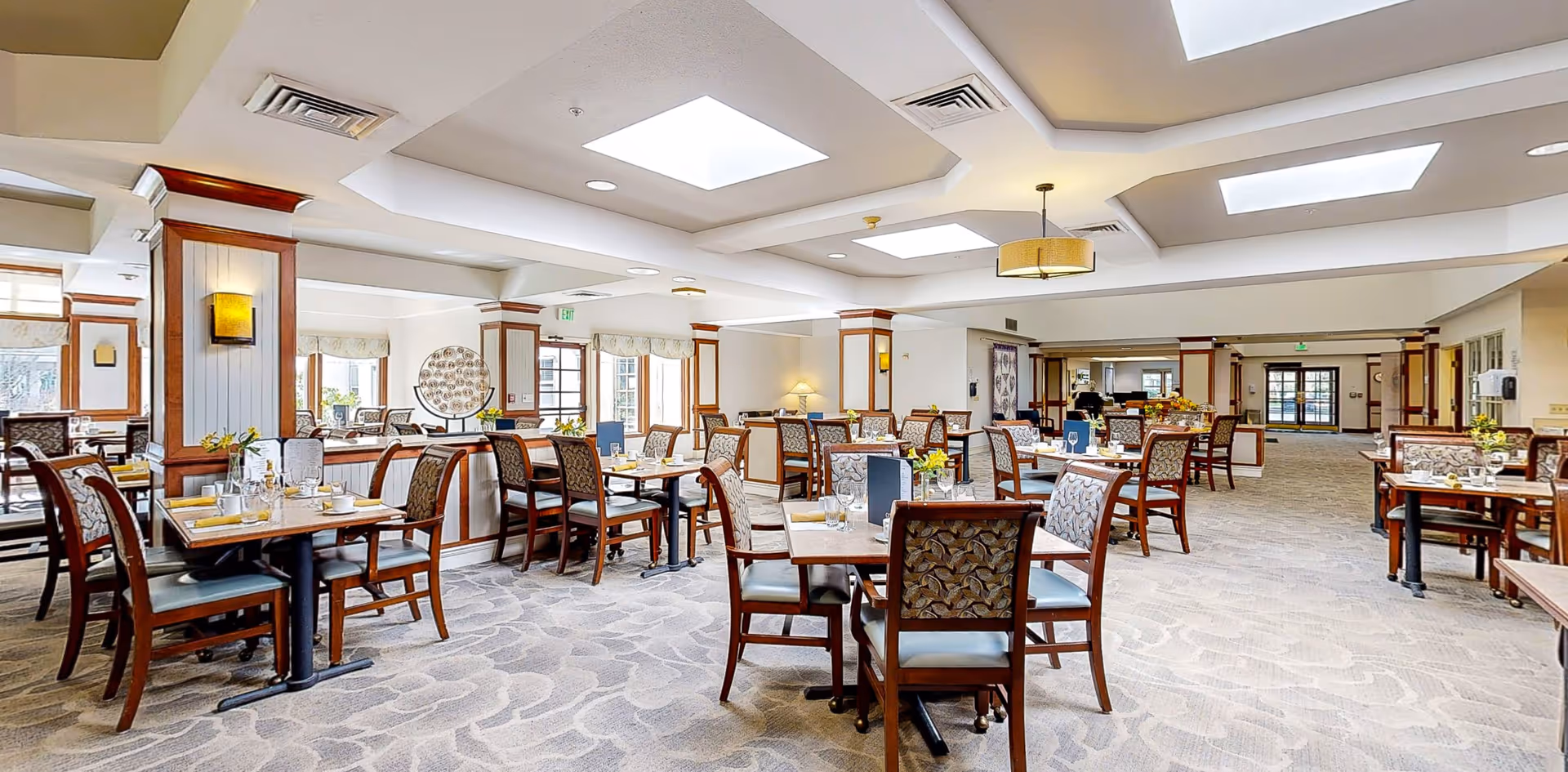 A spacious and well-lit dining room with multiple wooden tables and chairs arranged neatly. The room features large skylights in the ceiling, carpeted floors with a patterned design, and decorative columns with wood accents. Tables are set with placemats, glassware, and small flower arrangements, creating an inviting atmosphere.