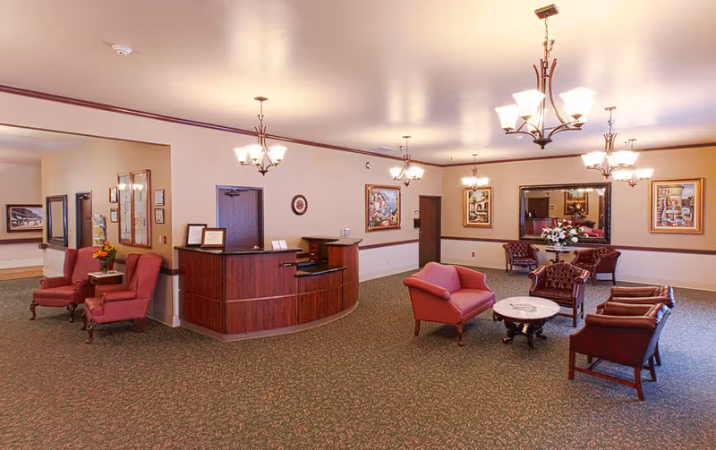 A spacious, well-lit common area with multiple seating arrangements including red upholstered chairs and sofas around small tables. There is a wooden reception desk in the middle, decorative paintings on the walls, and elegant ceiling light fixtures. The room has a carpeted floor and neutral-colored walls with a dark trim.