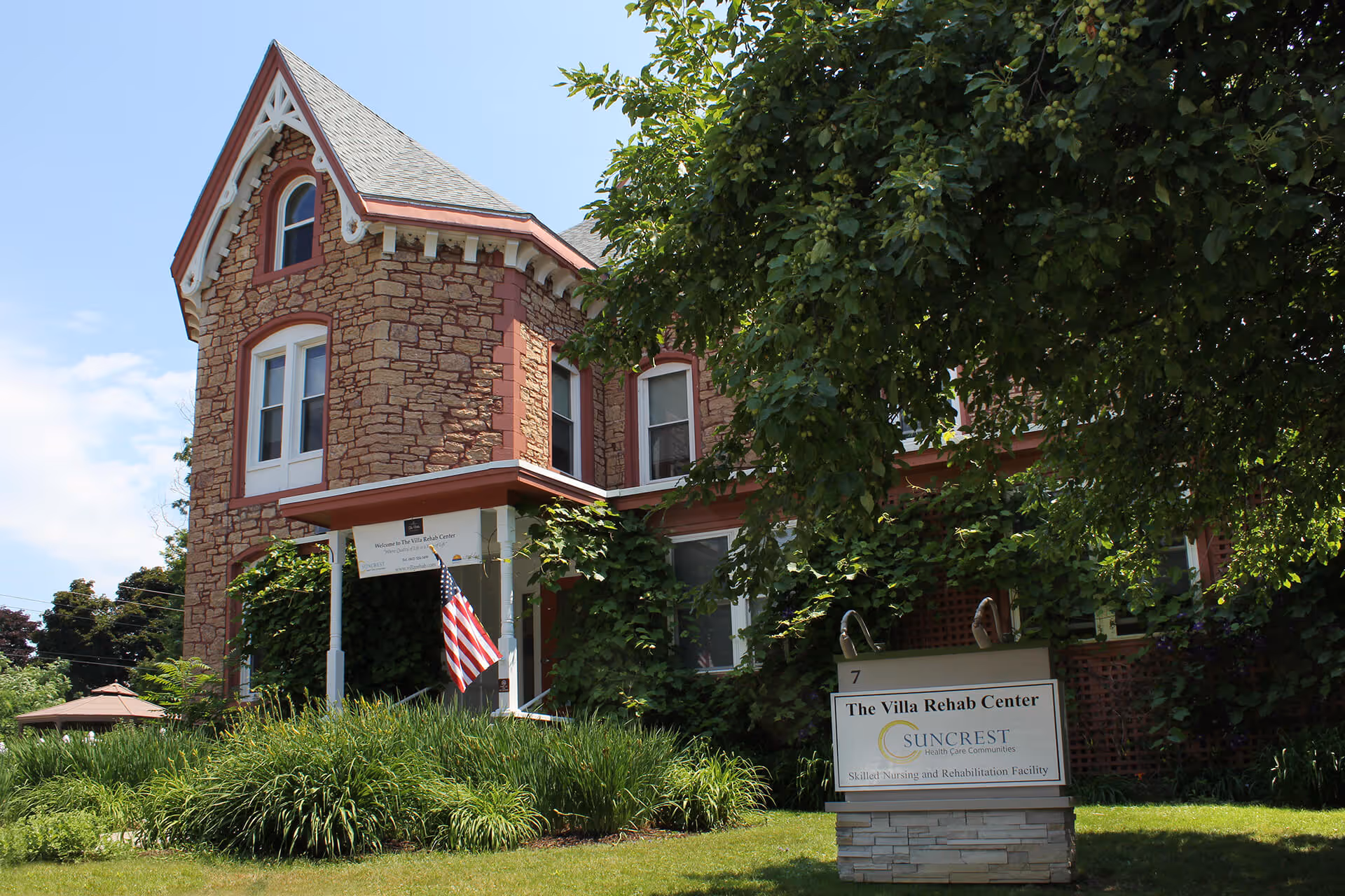 Exterior view of The Villa Rehab Center, a stone building with red trim and a steep roof, partially covered by green trees and bushes. An American flag hangs near the entrance, and a sign in front reads 'The Villa Rehab Center, Suncrest Health Care Communities, Skilled Nursing and Rehabilitation Facility.'
