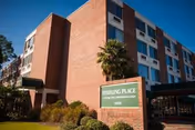 Exterior view of a multi-story brick building under a clear blue sky with a green sign in front that reads 'Sterling Place Assisted Living and Memory Care'. There are some shrubs and a small palm tree near the sign.