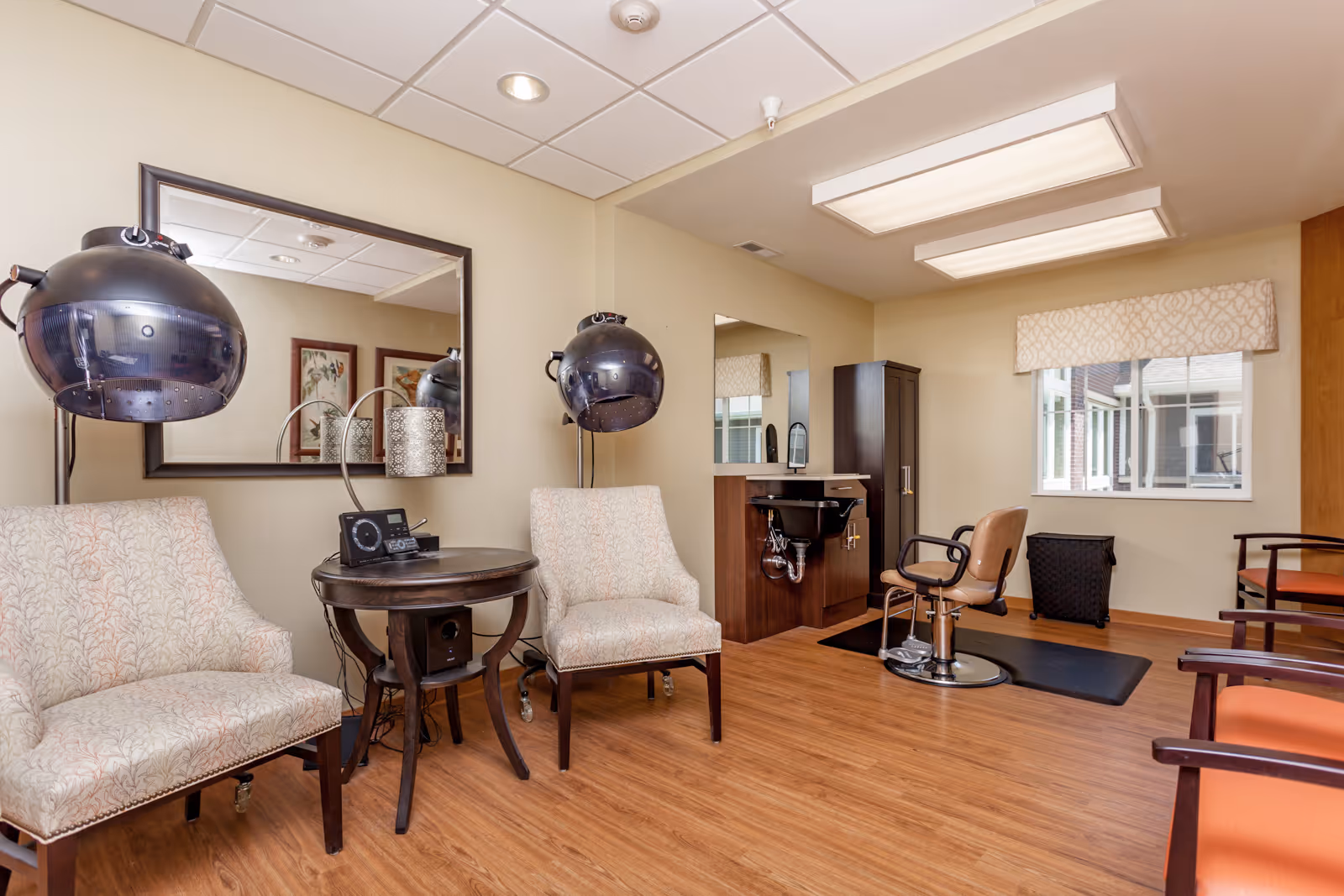 Interior view of a hair salon area in a senior living facility with two beige patterned chairs under black hair dryers, a round wooden table with a radio, a salon chair in front of a sink and mirror, wooden flooring, and a window with a patterned valance.