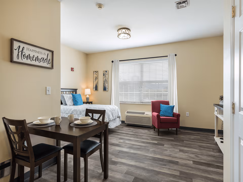 A bright and cozy studio apartment with a small dining table set for two, a bed with white bedding and blue accent pillows, a red armchair with a blue pillow, a window with white curtains and blinds, and light beige walls with a sign that reads 'Happiness is Homemade'.