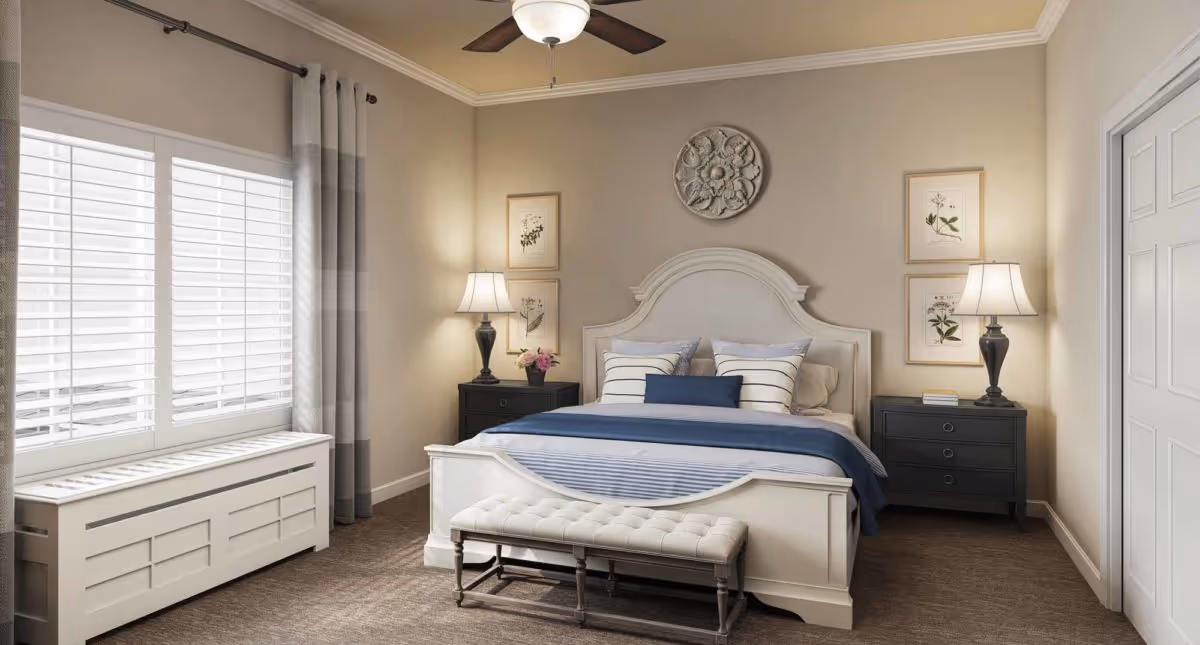 A well-lit bedroom with a white wooden bed frame, blue and white bedding, and a cushioned bench at the foot of the bed. There are two black nightstands on either side of the bed, each with a lamp and decorative items. The walls are beige with framed botanical prints and a decorative round wall piece above the headboard. A large window with white shutters and gray curtains is on the left side of the room.