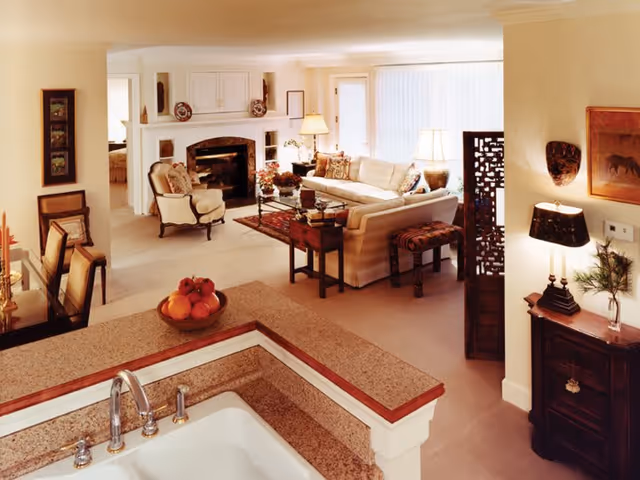 View of a cozy living room and dining area from the kitchen. The living room features a white sofa, armchair, coffee table, fireplace, and decorative items. The dining area has a table with chairs. The kitchen counter with a sink and a bowl of fruit is visible in the foreground. The room is warmly lit with lamps and natural light from a large window with vertical blinds.