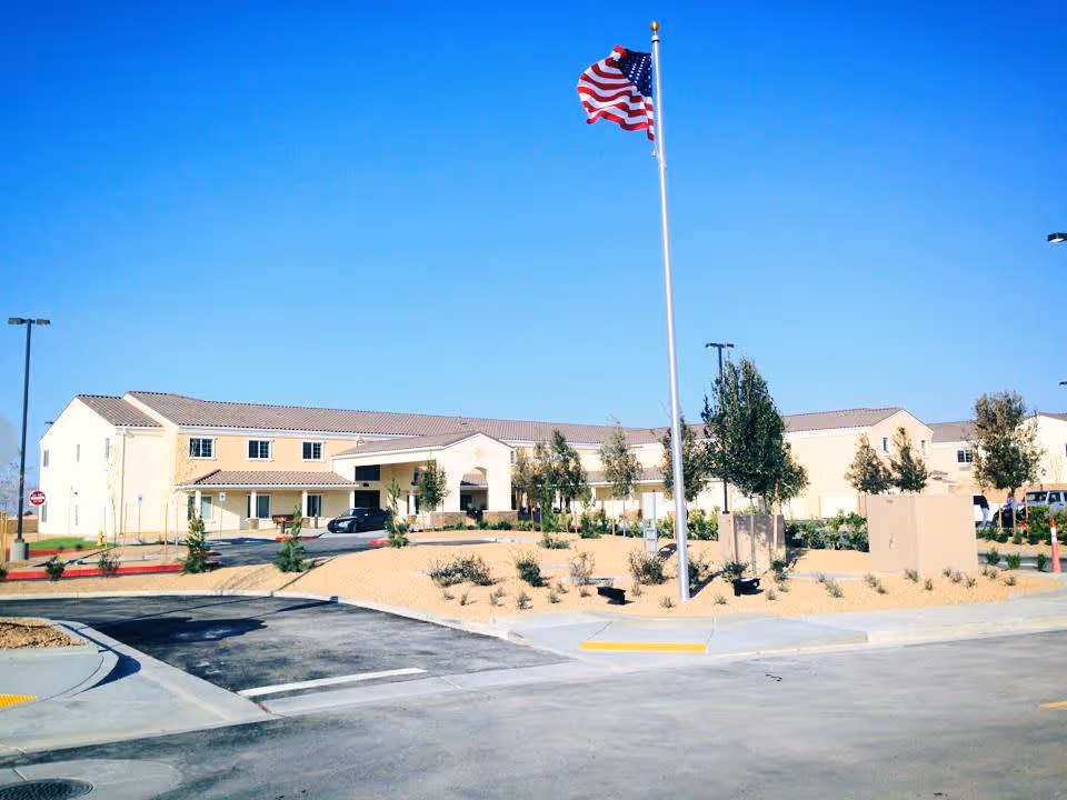 Exterior view of a large, beige senior living facility building under a clear blue sky with an American flag on a tall flagpole in the foreground, surrounded by landscaped areas with small trees and shrubs, and a paved driveway and parking area.