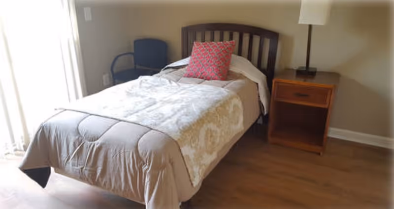 Small bedroom with a single bed with a patterned pillow, wooden nightstand and lamp, a chair, and a sliding glass door.
