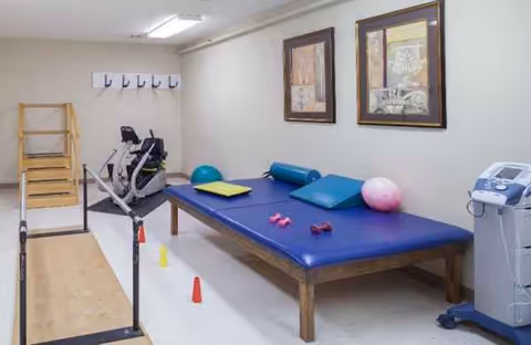 Therapy room with a padded table, parallel bars, exercise equipment, and balance balls.