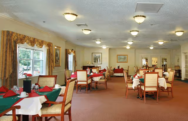 A spacious dining room with multiple tables covered in white and green tablecloths, each set with red napkins, cups, and glasses. The room has beige walls, framed artwork, large windows with patterned curtains, and ceiling lights providing warm illumination.