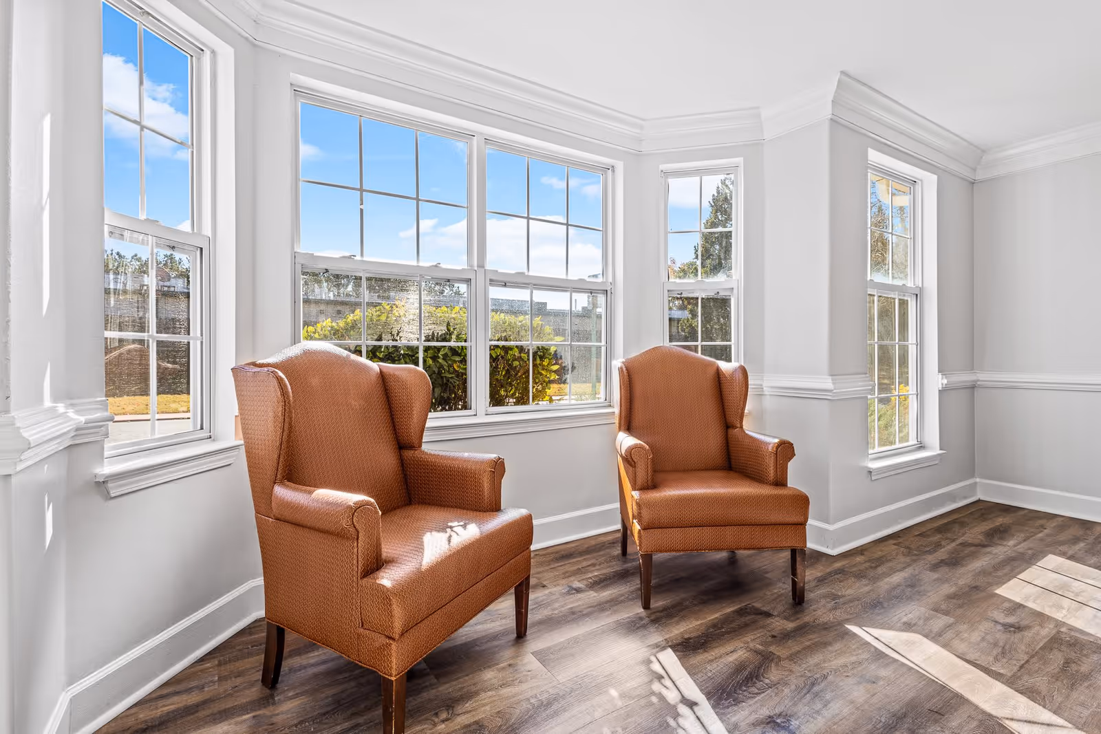 A bright room with large windows letting in natural light, featuring two brown upholstered armchairs on a wood floor with white walls and crown molding.
