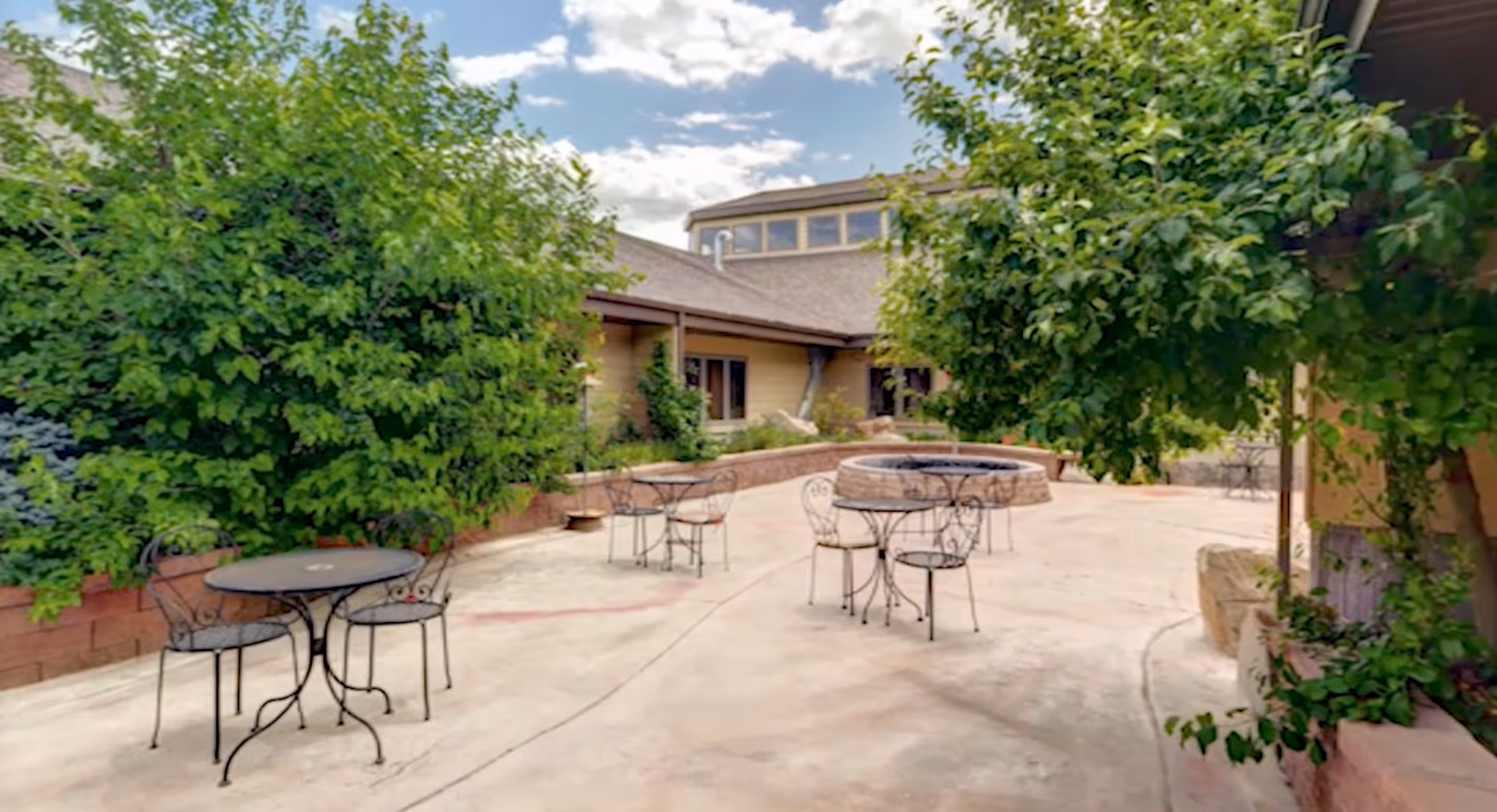 Outdoor patio area with several round metal tables and chairs, surrounded by green trees and bushes, with a building in the background under a partly cloudy sky.