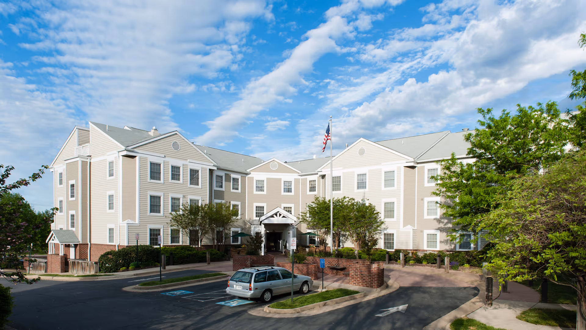 Exterior view of a three-story senior living facility building with beige siding and white trim under a partly cloudy blue sky. There is a flagpole with an American flag in front of the entrance, surrounded by trees and landscaping. A silver car is parked in a handicapped parking space near the entrance.