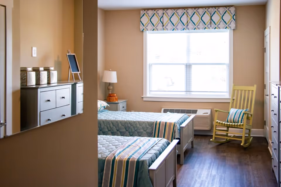 Twin bedroom with two beds, a bedside table and lamp, a window with a patterned valance, and a yellow rocking chair.