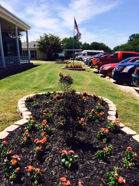 A landscaped garden bed with blooming pink flowers and a small shrub in the center, bordered by light-colored stones. The garden is situated on a grassy lawn next to a parking lot with several parked cars and a building with a covered porch on the left. An American flag is flying on a flagpole in the background under a partly cloudy blue sky.