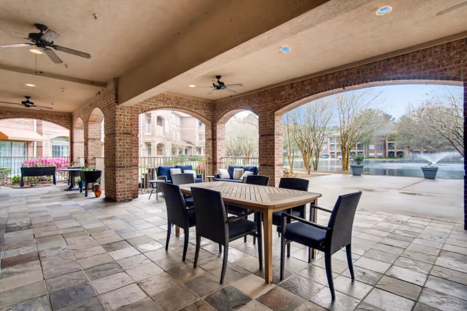 Covered outdoor patio area with a wooden table and six black chairs, ceiling fans, brick arches, and a view of a pond with fountains and trees in the background.