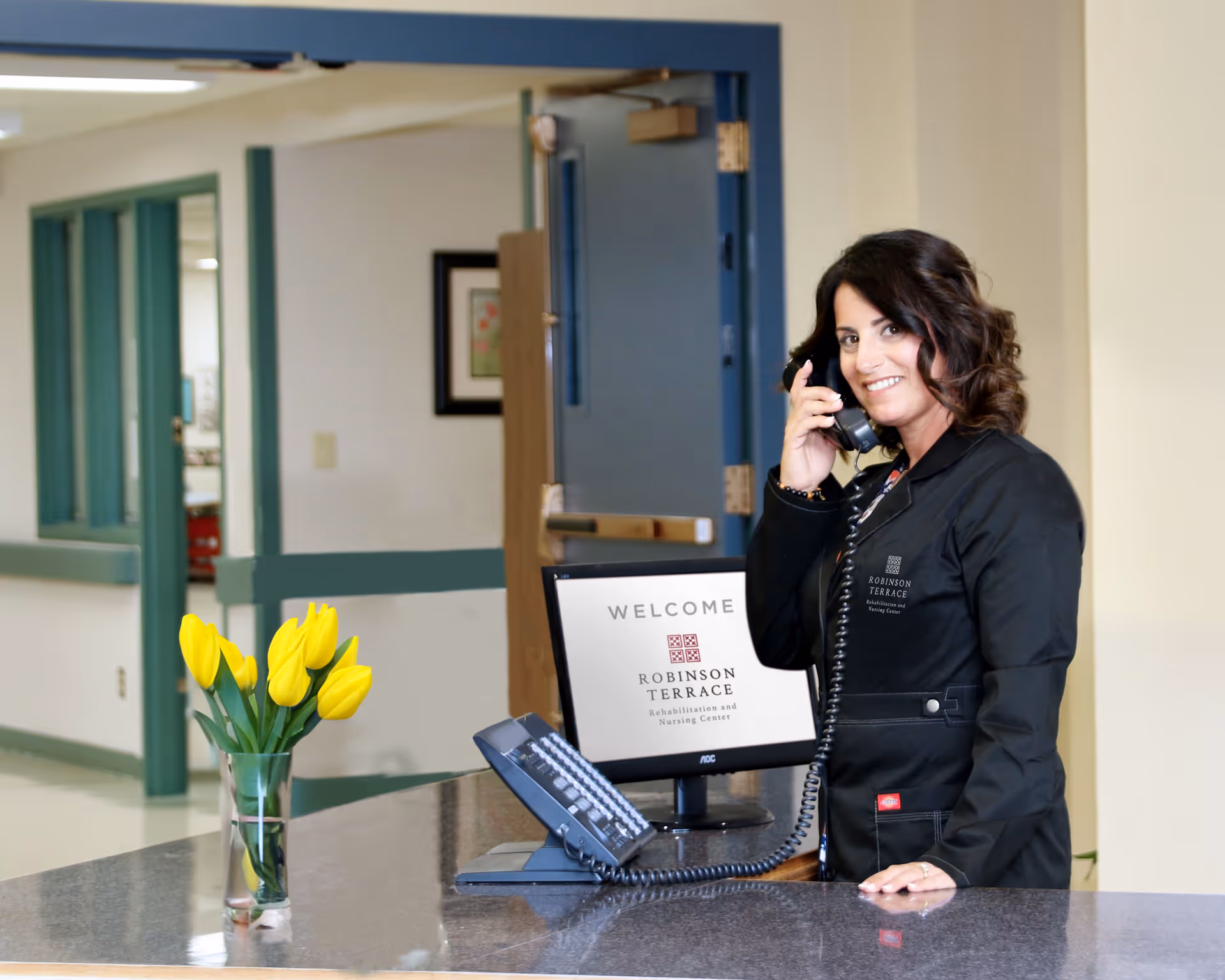 A woman wearing a black jacket with Robinson Terrace logo is standing behind a reception desk, smiling and talking on a corded telephone. On the desk is a vase with yellow tulips and a computer monitor displaying a welcome message for Robinson Terrace Rehabilitation and Nursing Center. The background shows a hallway with green trim and an open blue door.