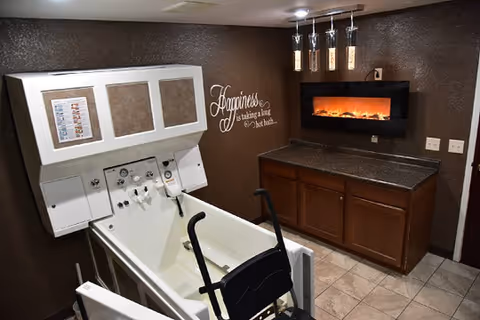 A bathroom designed for assisted bathing with a large white bathtub equipped with handrails and controls, a wheelchair positioned next to the tub, dark brown walls, a countertop with wooden cabinets, a modern electric fireplace mounted above the counter, and a wall decal that reads 'Happiness is taking a long hot bath.'