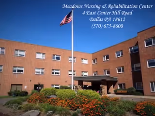 Exterior view of Meadows Nursing & Rehabilitation Center, a brick building with multiple windows, an American flag on a flagpole in front, and a landscaped garden with flowers and shrubs under a clear blue sky.