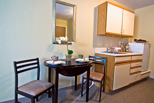 Small kitchenette and dining area with a round table set for two, two chairs, a wall mirror, sink, cabinets and a mini fridge.