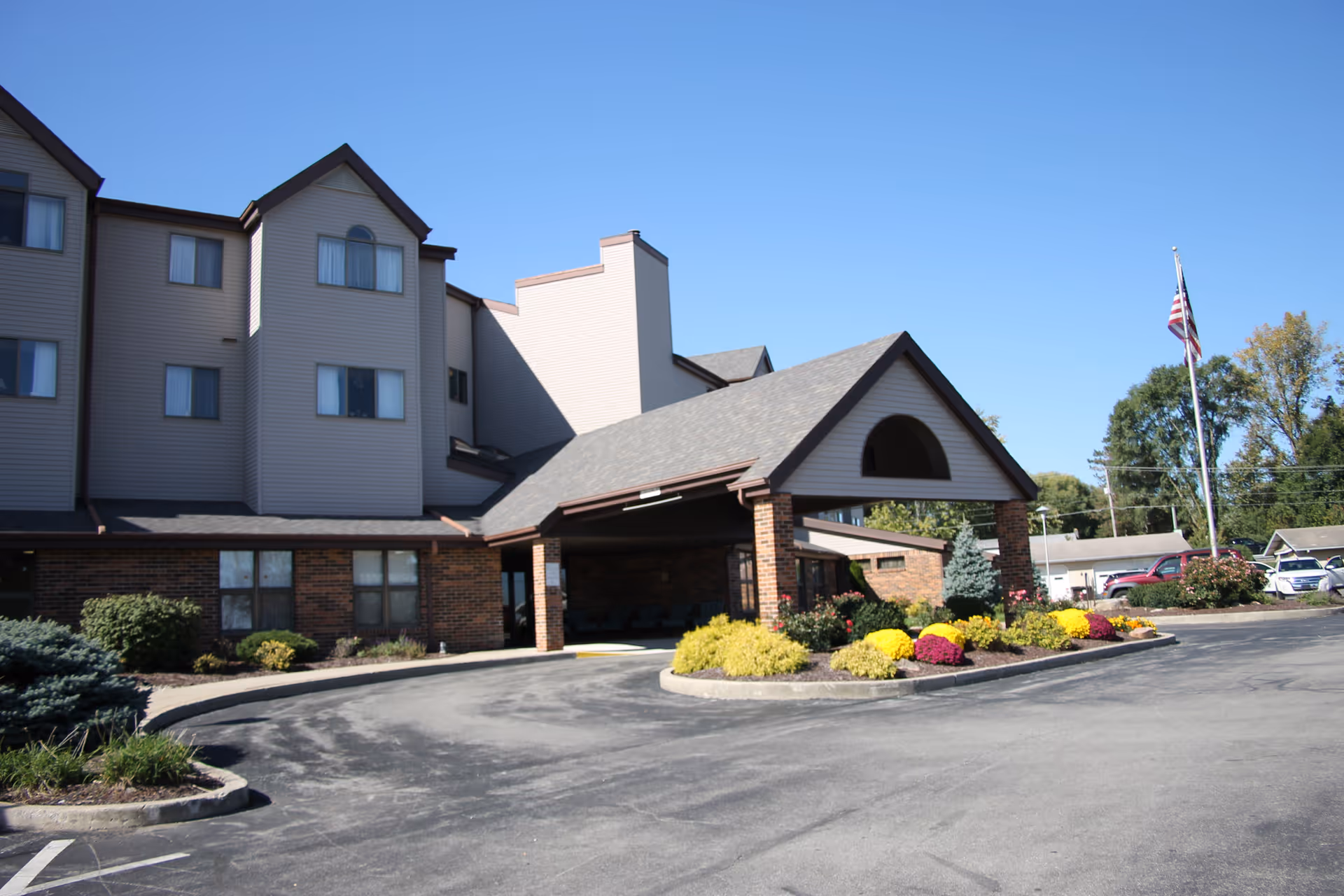 Exterior view of a multi-story senior living facility building with a covered entrance, landscaped flower beds, and an American flag on a flagpole under a clear blue sky.