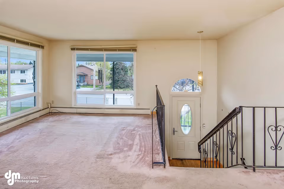 Carpeted front living room with large windows overlooking the street, a staircase and the entry door.