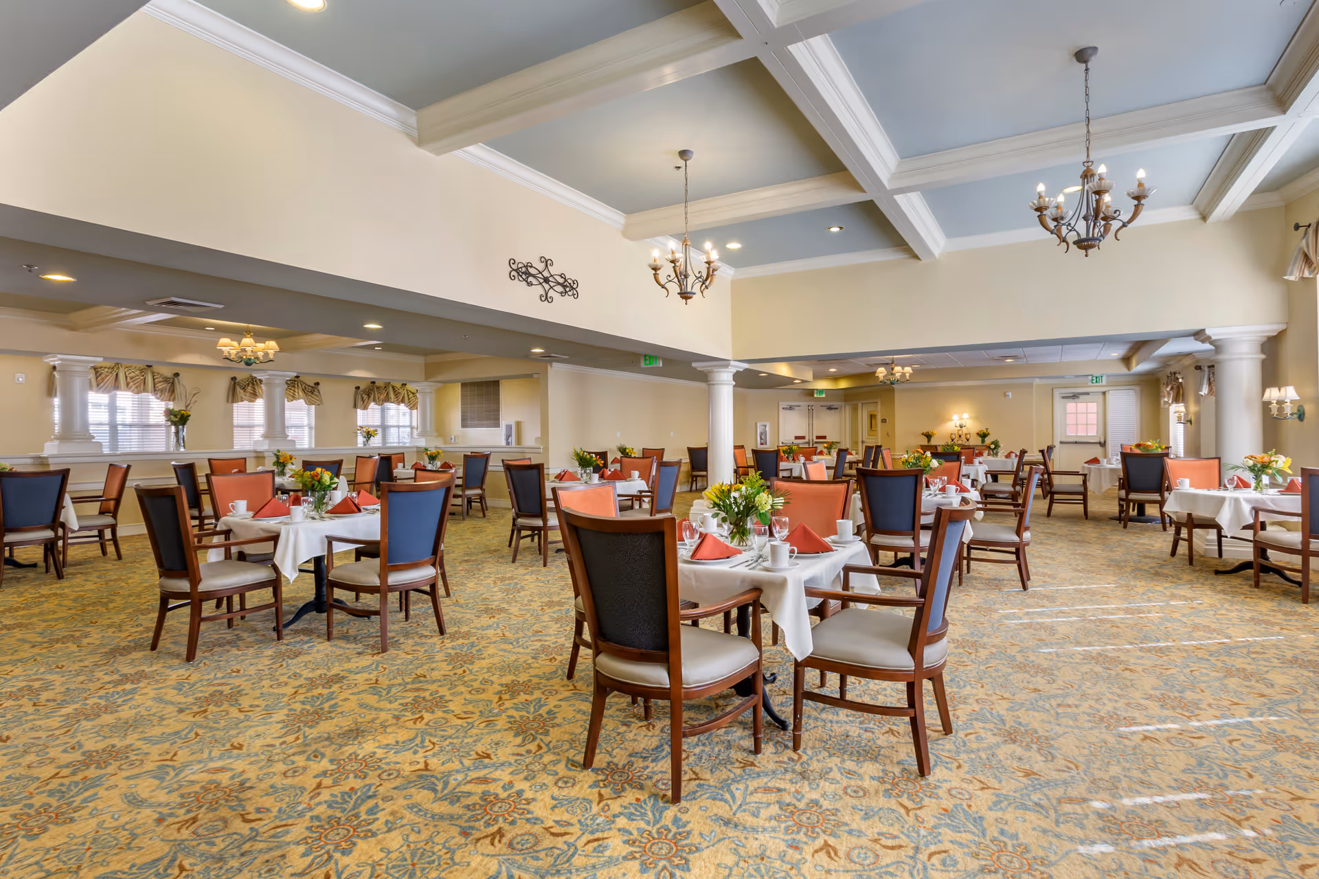 Spacious dining room with multiple set tables and chairs, chandeliers, and floral centerpieces.