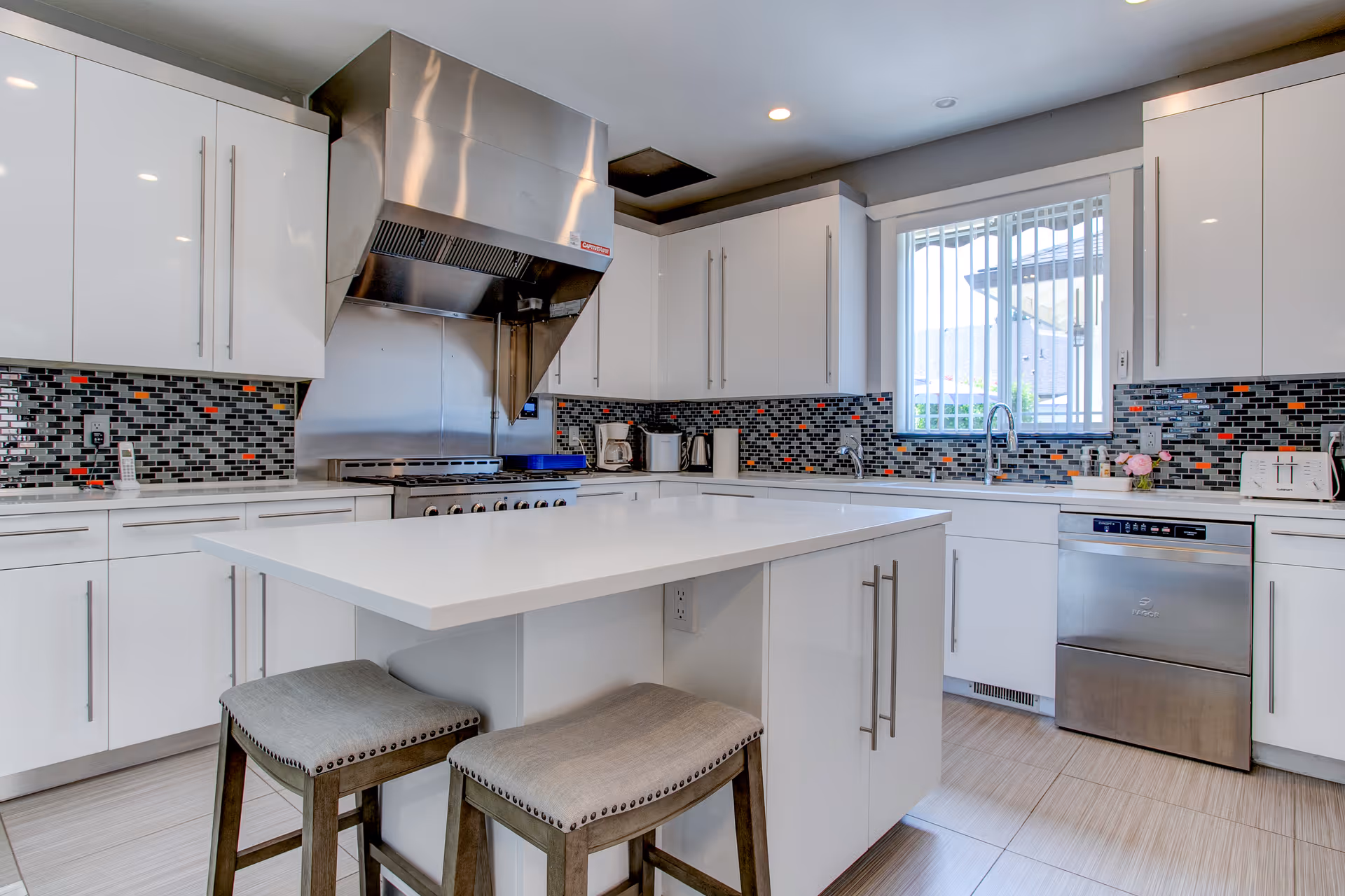 Modern kitchen with white cabinets, a large white island with two cushioned stools, stainless steel appliances including a dishwasher and a large range hood, and a window with vertical blinds above the sink. The backsplash features a mosaic tile pattern in black, gray, white, and orange.
