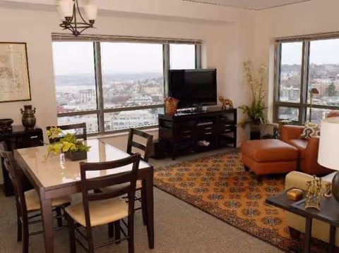 Bright living room with a dining table, sofa, TV, and city views through large windows.
