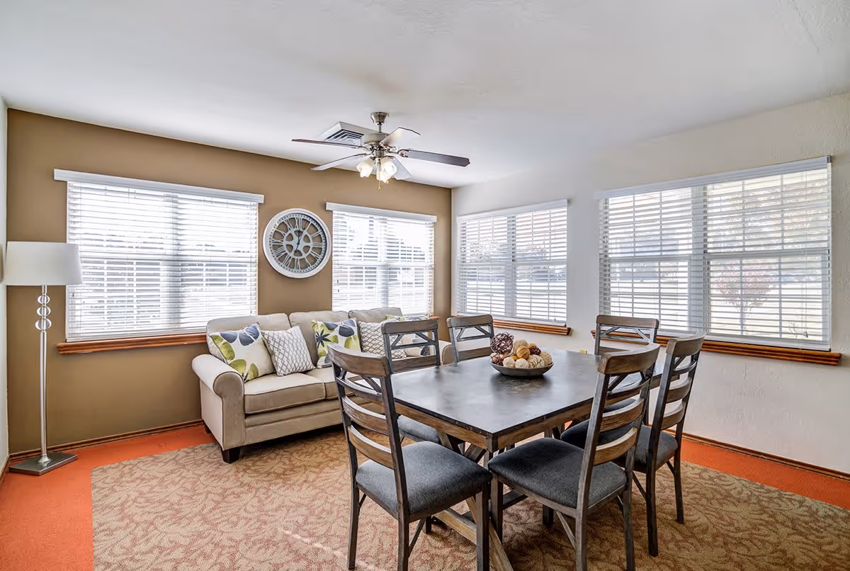 A bright room with multiple windows featuring white blinds. There is a beige sofa with patterned cushions against a brown accent wall, a large decorative clock above the sofa, and a floor lamp to the left. In the center of the room is a rectangular dining table with six wooden chairs, and a decorative bowl with assorted items on the table. The floor has a patterned carpet with an orange border.
